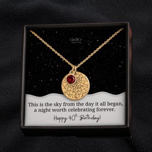 May include: A gold-coloured necklace with a circular pendant, a red gemstone, and a celestial map design. The necklace is in a black box with the text: "This is the sky from the day it all began, a night worth celebrating forever. Happy 40th Birthday!"