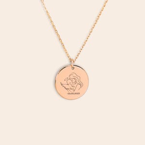 K&ouml;nnte beinhalten: Goldkette mit einem runden Anh&auml;nger, der eine Linienzeichnung einer Mutter zeigt, die ein Baby mit Engelsfl&uuml;geln h&auml;lt. Der Anh&auml;nger ist mit dem Datum "03.05.2023" graviert.