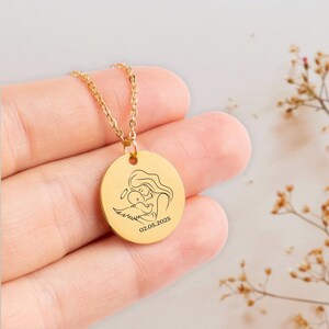 K&ouml;nnte beinhalten: Goldkette mit einem runden Anh&auml;nger, der eine Linienzeichnung einer Mutter zeigt, die ein Baby mit Engelsfl&uuml;geln h&auml;lt. Der Anh&auml;nger ist mit dem Datum 02.05.2025 graviert.