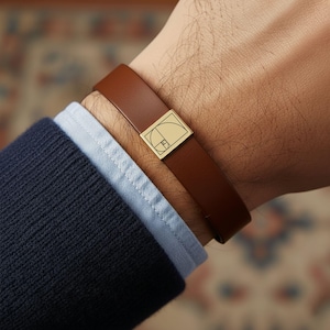 May include: A brown leather bracelet with a gold-colored rectangular clasp featuring a geometric design. The bracelet is worn on a wrist, paired with a navy blue sweater and a light blue collared shirt.