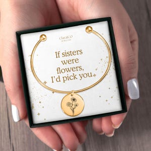 May include: Gold bangle bracelet with a charm featuring a floral design. The bracelet comes in a box with the text "If sisters were flowers, I'd pick you."