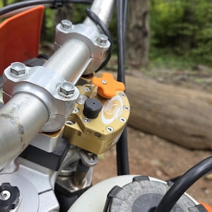 May include: Close-up of a motorcycle handlebar assembly. The silver handlebar is secured with silver bolts. A gold and black component with an orange adjustment knob is visible. The motorcycle has an orange fairing and a white fuel tank.
