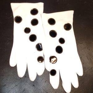 May include: A pair of white gloves adorned with large, black, circular embellishments. The gloves are made of a smooth material and feature a classic design. The black circles are evenly spaced across the surface of the gloves.