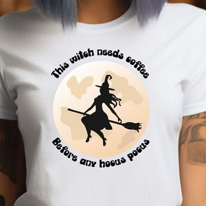 Op de afbeelding: Wit T-shirt met een afbeelding van een heks op een bezem voor een maan. De tekst op het shirt luidt: "This witch needs coffee before any hocus pocus."