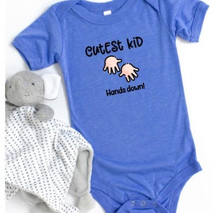 K&ouml;nnte beinhalten: Ein blauer Baby-Body mit dem Text "Cutest Kid Hands down!" in Schwarz. Ein graues Stofftier und eine wei&szlig;-grau gemusterte Decke liegen neben dem Body.