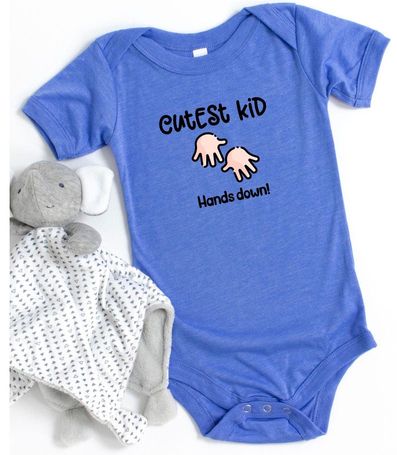 K&ouml;nnte beinhalten: Ein blauer Baby-Body mit dem Text "Cutest Kid Hands down!" in Schwarz. Ein graues Stofftier und eine wei&szlig;-grau gemusterte Decke liegen neben dem Body.