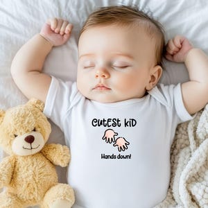K&ouml;nnte beinhalten: Ein schlafendes Baby in einem wei&szlig;en Body mit dem Text "Cutest kid Hands down!" und einer Cartoon-Handgrafik. Ein hellbrauner Teddyb&auml;r liegt neben dem Baby auf einem wei&szlig;en Laken.