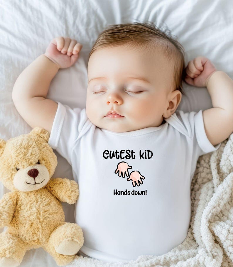 K&ouml;nnte beinhalten: Ein schlafendes Baby in einem wei&szlig;en Body mit dem Text "Cutest kid Hands down!" und einer Cartoon-Handgrafik. Ein hellbrauner Teddyb&auml;r liegt neben dem Baby auf einem wei&szlig;en Laken.