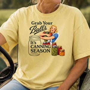 Pode incluir: Uma t-shirt de manga curta amarelo-pálido com um gráfico de uma pessoa a segurar frascos de conservas. O texto diz Grab Your Balls It's CANNING SEASON. A ilustração inclui vários frascos de vidro com vegetais verdes e cor de laranja.
