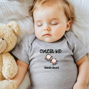K&ouml;nnte beinhalten: Ein schlafendes Baby in einem grauen Body mit dem Text "Cutest Kid Hands down!" und einer Cartoon-Handgrafik. Ein beiger Teddyb&auml;r liegt neben dem Baby auf einem wei&szlig;en Laken.