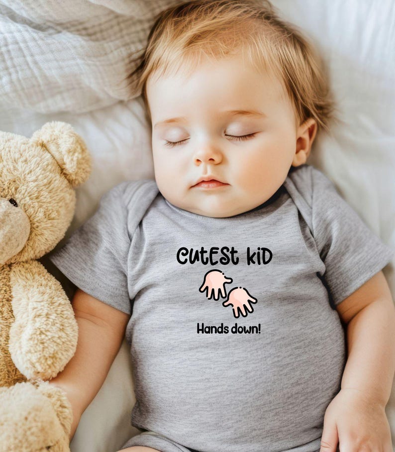 K&ouml;nnte beinhalten: Ein schlafendes Baby in einem grauen Body mit dem Text "Cutest Kid Hands down!" und einer Cartoon-Handgrafik. Ein beiger Teddyb&auml;r liegt neben dem Baby auf einem wei&szlig;en Laken.