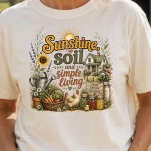 May include: A cream-coloured t-shirt featuring a graphic print with the text Sunshine, soil, and simple living. The design includes a farmhouse, a sunflower, a watering can, a basket of vegetables, a chicken, and jars labelled Grow Gather Grateful.