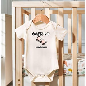 K&ouml;nnte beinhalten: Cremefarbener Baby-Body mit dem Text "Cutest Kid" &uuml;ber einer Grafik von zwei H&auml;nden und dem Satz "Hands down!" Der Body h&auml;ngt an einem Holzkleiderb&uuml;gel vor einem Holzbettchen.