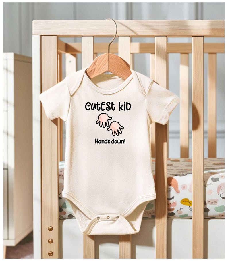 K&ouml;nnte beinhalten: Cremefarbener Baby-Body mit dem Text "Cutest Kid" &uuml;ber einer Grafik von zwei H&auml;nden und dem Satz "Hands down!" Der Body h&auml;ngt an einem Holzkleiderb&uuml;gel vor einem Holzbettchen.