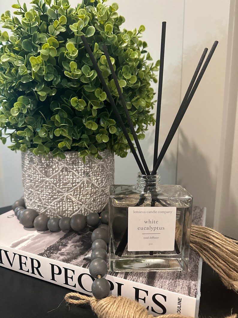 May include: A clear glass bottle with a white label that reads "lemieux candle company white eucalyptus reed diffuser". The bottle is filled with a clear liquid and has black reeds sticking out of the top. The bottle is sitting on a book with a gray and white pattern. There are gray beads and a piece of twine on the book.