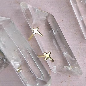 May include: Clear quartz crystal rings with gold-colored cross accents. The rings are set on clear quartz crystals, with a light pink background. The crystals are irregularly shaped and have a natural, raw appearance.