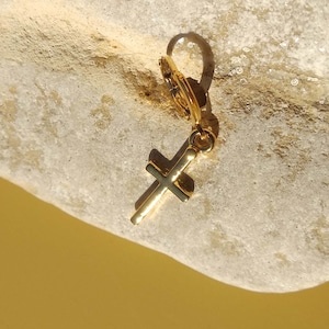 May include: Gold cross charm earring with a hoop style closure. The earring is on a light brown textured surface.