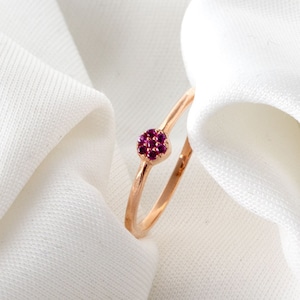 Puede incluir: Un delicado anillo de oro rosa con un pequeño grupo de piedras de color rojo rubí engastadas en el centro.