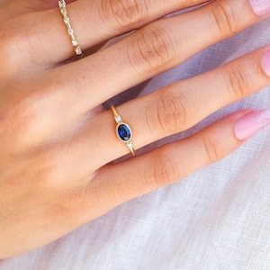 May include: Two gold rings on a hand. The ring on the index finger has a blue oval gemstone surrounded by smaller clear gemstones. The ring on the middle finger has a row of clear gemstones.