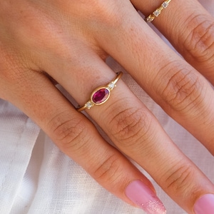 Puede incluir: Una mano con dos anillos de oro. El anillo en el dedo índice tiene una sola piedra preciosa rosa de forma ovalada rodeada de piedras preciosas transparentes más pequeñas. El anillo en el dedo medio es una banda delgada con piedras preciosas transparentes. La mano tiene esmalte de uñas rosa con brillo en las puntas.