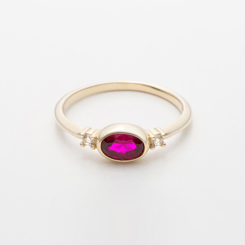 May include: A gold ring with a large oval-shaped ruby gemstone and two smaller round clear gemstones on either side.