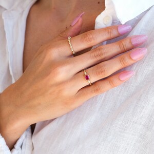 May include: A close-up of a hand wearing two gold rings. The ring on the index finger has a small oval ruby gemstone surrounded by tiny diamonds. The ring on the middle finger is a delicate band of small diamonds.