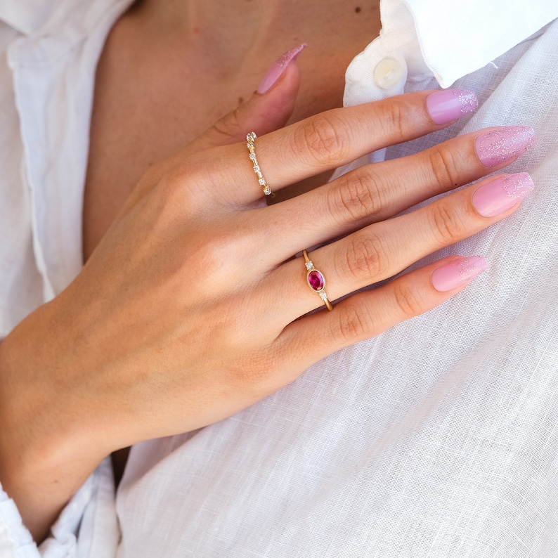 May include: A close-up of a hand wearing two gold rings. The ring on the index finger has a small oval ruby gemstone surrounded by tiny diamonds. The ring on the middle finger is a delicate band of small diamonds.