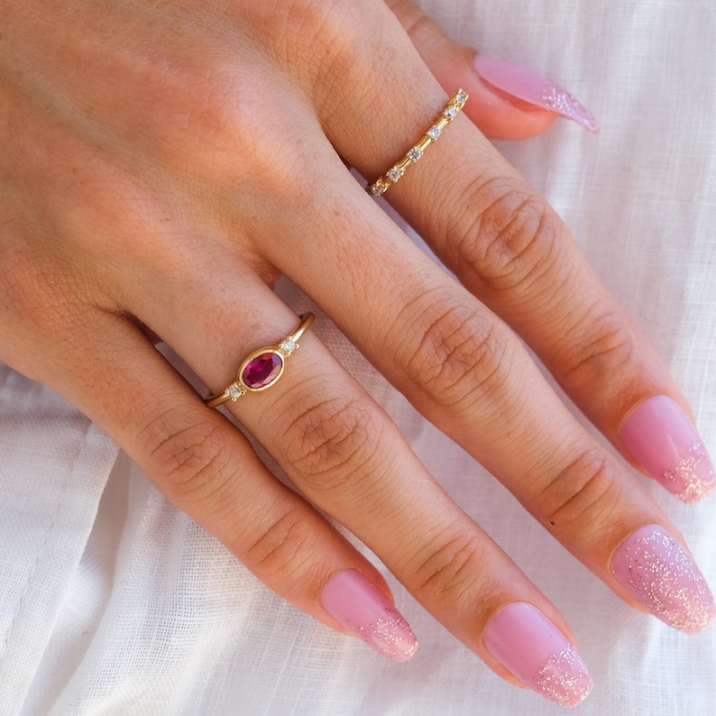 May include: A hand wearing two gold rings. The ring on the index finger has a single oval-shaped pink gemstone surrounded by smaller clear gemstones. The ring on the middle finger is a thin band with clear gemstones. The hand has pink nail polish with glitter on the tips.