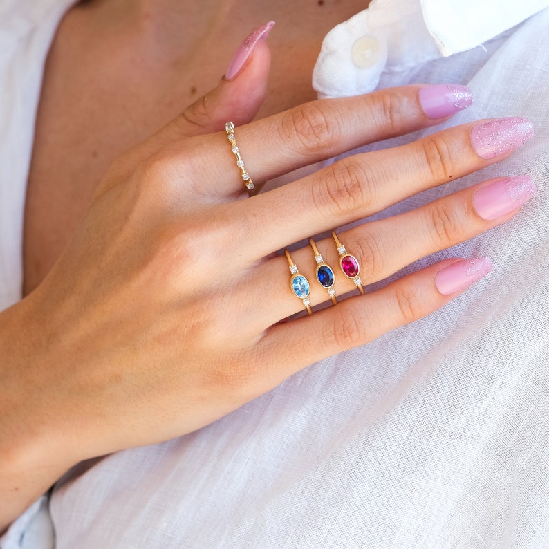 May include: A hand wearing four gold rings with oval-shaped gemstones. The rings feature a blue topaz, a sapphire, a ruby, and a diamond band.