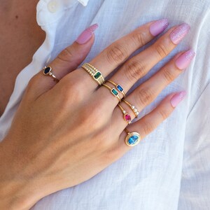 May include: A hand wearing a variety of gold rings with different colored gemstones, including a blue oval stone, a pink stone, a green stone, and a blue rectangular stone.