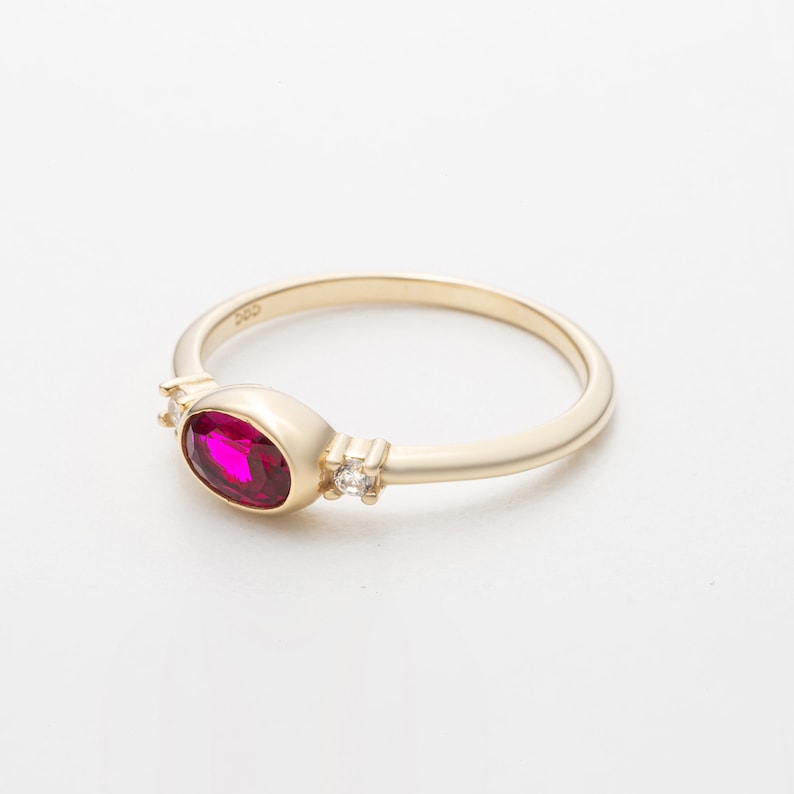 May include: A gold ring with a single oval-shaped ruby gemstone and two smaller clear gemstones. The ring is set on a white background.