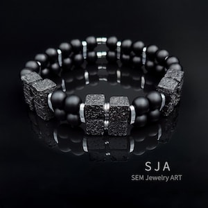 K&ouml;nnte beinhalten: Ein schwarzes Armband mit strukturierten quadratischen Elementen und silberfarbenen Metallringen. Auf dem Armband steht der Text "SJA SEM Jewelry ART". Das Armband wird auf einer reflektierenden schwarzen Oberfl&auml;che pr&auml;sentiert.