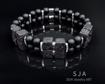 Peut inclure: Un bracelet noir composé de perles et d'éléments carrés texturés, avec des anneaux en métal argenté. Le bracelet porte le texte "SJA SEM Jewelry ART". Il est présenté sur une surface noire réfléchissante.