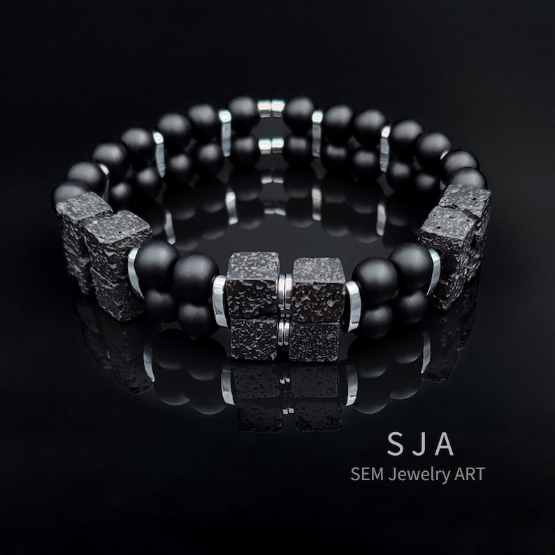 K&ouml;nnte beinhalten: Ein schwarzes Armband mit strukturierten quadratischen Elementen und silberfarbenen Metallringen. Auf dem Armband steht der Text "SJA SEM Jewelry ART". Das Armband wird auf einer reflektierenden schwarzen Oberfl&auml;che pr&auml;sentiert.