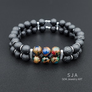 Könnte beinhalten: Ein zweireihiges Armband mit schwarzen Lavaperlen und bunten, gemusterten Edelsteinperlen. Das Armband wird durch silberfarbene Metallstäbe akzentuiert. Der Text "SJA SEM Jewelry Art" ist sichtbar.