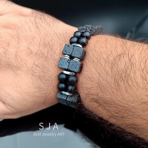 K&ouml;nnte beinhalten: Armband aus schwarzem Lavastein und Onyxperlen mit silberfarbenen Metallakzenten. Das Armband hat rechteckige Lavasteinperlen und runde Onyxperlen, die durch silberfarbene Metallringe verbunden sind. Das Armband befindet sich an einem Handgelenk.