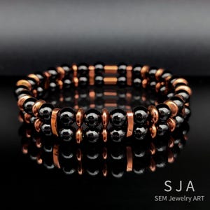 May include: Two-strand bracelet featuring black, spherical beads and copper-colored cylindrical and circular accents. The bracelet is displayed on a reflective black surface. The text "SJA SEM Jewelry ART" is visible in the lower right corner.