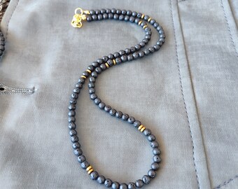 Könnte beinhalten: Eine schwarze Lava-Perlenkette mit goldenen Akzenten. Die Kette liegt auf einem grauen Stoffhintergrund.
