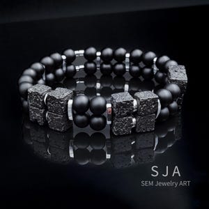 K&ouml;nnte beinhalten: Ein schwarzes Perlenarmband mit quadratischen, strukturierten Akzenten und silberfarbenen Metalldetails. Das Armband hat ein zweireihiges Design mit mattschwarzen Perlen. Der Text "SJA SEM Jewelry Art" ist sichtbar.