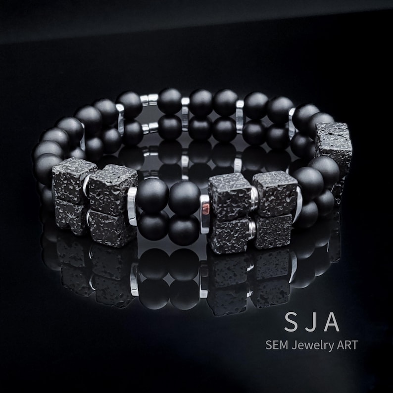 K&ouml;nnte beinhalten: Ein schwarzes Perlenarmband mit quadratischen, strukturierten Akzenten und silberfarbenen Metalldetails. Das Armband hat ein zweireihiges Design mit mattschwarzen Perlen. Der Text "SJA SEM Jewelry Art" ist sichtbar.