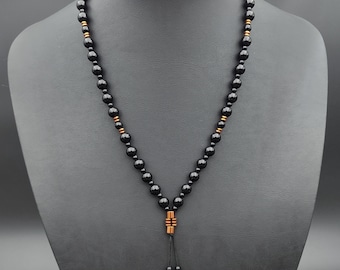 May include: A black beaded necklace with copper-coloured accents and a tassel pendant. The necklace is displayed on a black mannequin. The beads are round and evenly spaced. The pendant has a rectangular design. The necklace is a piece of jewellery.