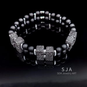 K&ouml;nnte beinhalten: Ein schwarzes Perlenarmband mit strukturierten, quadratischen Akzenten und silberfarbenen Abstandshaltern. Das Armband hat matte schwarze Perlen und wird auf einer reflektierenden schwarzen Oberfl&auml;che pr&auml;sentiert. Der Text "SJA SEM Jewelry ART" ist unten rechts sichtbar.