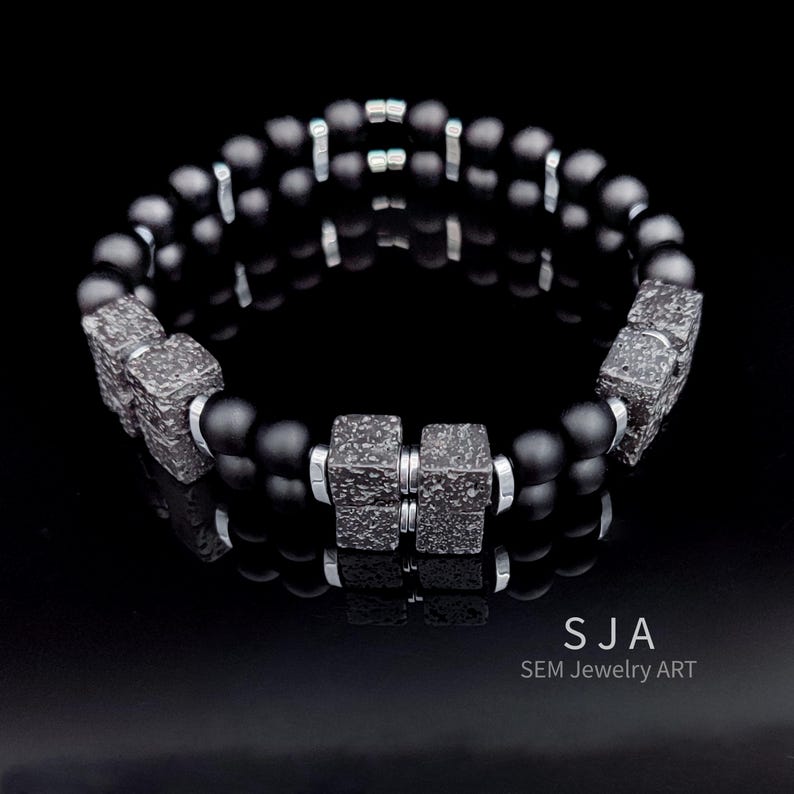 K&ouml;nnte beinhalten: Ein schwarzes Perlenarmband mit strukturierten, quadratischen Akzenten und silberfarbenen Abstandshaltern. Das Armband hat matte schwarze Perlen und wird auf einer reflektierenden schwarzen Oberfl&auml;che pr&auml;sentiert. Der Text "SJA SEM Jewelry ART" ist unten rechts sichtbar.