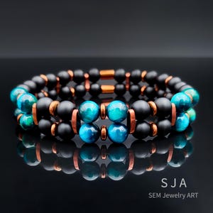 Könnte beinhalten: Zwei gestapelte Armbänder mit schwarzen matten Perlen und leuchtend blau-grünen Tigeraugenperlen. Kupferfarbene Metallakzente und Verbinder setzen Kontraste. Die Armbänder werden auf einer reflektierenden schwarzen Oberfläche präsentiert. Der Text "SJA SEM Jewelry ART" ist sichtbar.