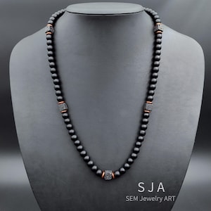 May include: A black beaded necklace with square, textured accents and copper-coloured spacers. The necklace is displayed on a black mannequin. The text "SJA SEM Jewelry ART" is visible at the bottom.