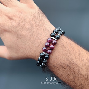 Könnte beinhalten: Doppelreihiges Armband mit schwarzen und burgunderfarbenen Perlen, getrennt durch silberfarbene Abstandshalter. Das Armband wird am Handgelenk getragen, das SJA SEM Jewelry ART Logo ist unten sichtbar.