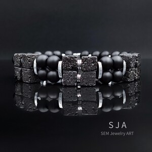 K&ouml;nnte beinhalten: Zwei schwarze Perlenarmb&auml;nder mit quadratischen, strukturierten schwarzen Steinakzenten und silberfarbenen Metalldetails. Die Armb&auml;nder werden auf einer reflektierenden schwarzen Oberfl&auml;che pr&auml;sentiert, die das Design des Schmucks hervorhebt. Der Text "SJA SEM Jewelry ART" ist enthalten.