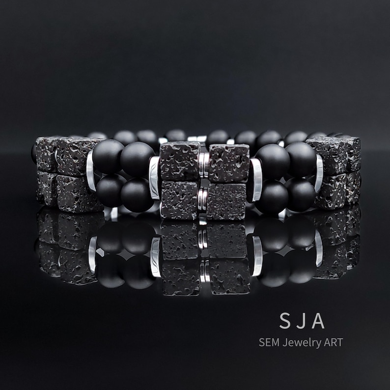 K&ouml;nnte beinhalten: Zwei schwarze Perlenarmb&auml;nder mit quadratischen, strukturierten schwarzen Steinakzenten und silberfarbenen Metalldetails. Die Armb&auml;nder werden auf einer reflektierenden schwarzen Oberfl&auml;che pr&auml;sentiert, die das Design des Schmucks hervorhebt. Der Text "SJA SEM Jewelry ART" ist enthalten.