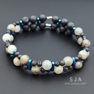 May include: A double-strand bracelet featuring black lava rock beads, light-colored gemstone beads, and blue metal accents. The bracelet is connected with a silver clasp. The jewelry is displayed on a dark surface.