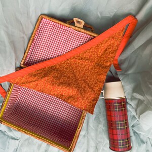 Puede incluir: Una cesta de picnic de mimbre con un forro a cuadros rojo y blanco. Una tela roja con círculos naranjas está doblada dentro de la cesta. Un termo rojo y negro a cuadros está sobre el fondo de tela blanca.